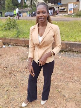 A woman is standing outdoors on a dirt and grass area, wearing a beige blazer over a white top, black pants, and cream-colored shoes. She has braided hair and is smiling at the camera. In the background, there are trees, a parking lot with a car, and several people gathered near a building.
