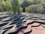 A wavy, abstract wooden deck structure is surrounded by lush green trees. The design features a combination of curved and angular lines, creating a visually striking pattern. Sunlight filters through the tree canopy, casting shadows and adding depth to the scene.
