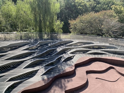 A wavy, abstract wooden deck structure is surrounded by lush green trees. The design features a combination of curved and angular lines, creating a visually striking pattern. Sunlight filters through the tree canopy, casting shadows and adding depth to the scene.