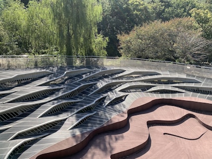 A wavy, abstract wooden deck structure is surrounded by lush green trees. The design features a combination of curved and angular lines, creating a visually striking pattern. Sunlight filters through the tree canopy, casting shadows and adding depth to the scene.