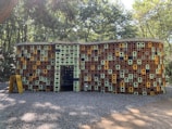 A structure made of stacked, colorful plastic crates arranged to form a wall with a wavy pattern. The crates are primarily brown, green, and yellow, creating a mosaic effect. The structure stands on a gravel path surrounded by trees and foliage, and a yellow A-frame sign is visible in the foreground.