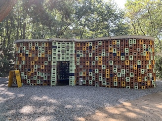 A structure made of stacked, colorful plastic crates arranged to form a wall with a wavy pattern. The crates are primarily brown, green, and yellow, creating a mosaic effect. The structure stands on a gravel path surrounded by trees and foliage, and a yellow A-frame sign is visible in the foreground.