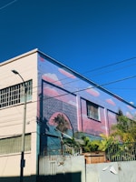 A building wall adorned with colorful mural art depicting palm trees and a sunset, creating a vibrant and tropical atmosphere. The building is surrounded by wire fences and some actual palm trees, enhancing the artistic theme.