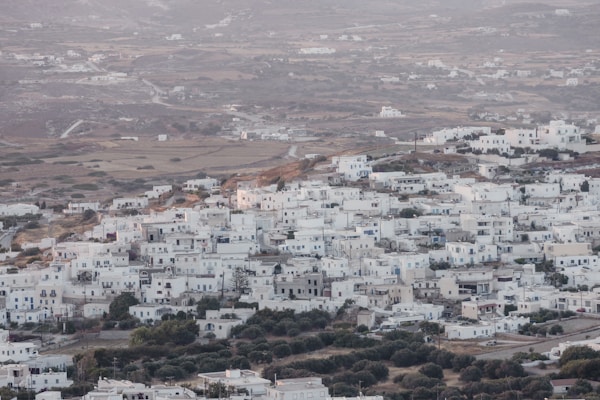 A sprawling town of white buildings situated on a gently rolling landscape. The architecture is uniform and traditional, with a notable absence of high-rise structures. The setting is rural, with patches of greenery and rustic terrain surrounding the town.