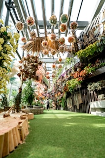 A lush indoor garden with a pathway covered in green artificial grass. The area is adorned with hanging floral arrangements in various colors, natural wood decor, and dense greenery on the walls. The space is brightly lit with natural light coming through a glass ceiling. Two people can be seen walking along the path.