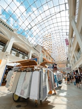 An indoor shopping mall with a high glass ceiling, allowing natural light to illuminate the space. The ceiling is decorated with colorful hanging orbs. Below, a display cart presents a variety of fabrics and textiles on racks, with people walking around, enjoying the shopping area.
