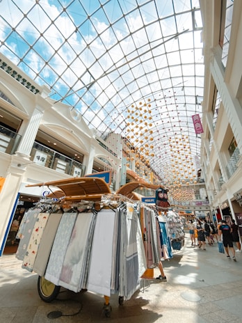 An indoor shopping mall with a high glass ceiling, allowing natural light to illuminate the space. The ceiling is decorated with colorful hanging orbs. Below, a display cart presents a variety of fabrics and textiles on racks, with people walking around, enjoying the shopping area.