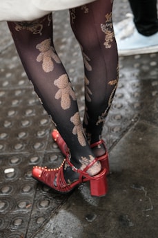 A pair of legs wearing decorative black tights with a teddy bear pattern. The person is also wearing strappy red high-heeled shoes. The ground is wet, reflecting a rainy environment with metal-textured flooring.