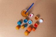 Several painted pine cones are arranged on a brown cardboard surface, accompanied by small cups containing colorful paint. A brush is immersed in a glass of orange liquid, suggesting a creative painting setup.