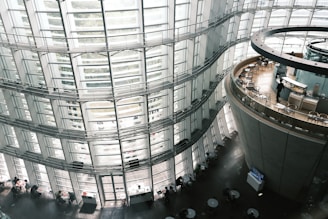 The interior of a modern architectural structure, featuring floor-to-ceiling curved glass windows and multiple levels. Tables and chairs are arranged in circular formations, and a circular café area is situated on the right. The space appears open and filled with natural light.