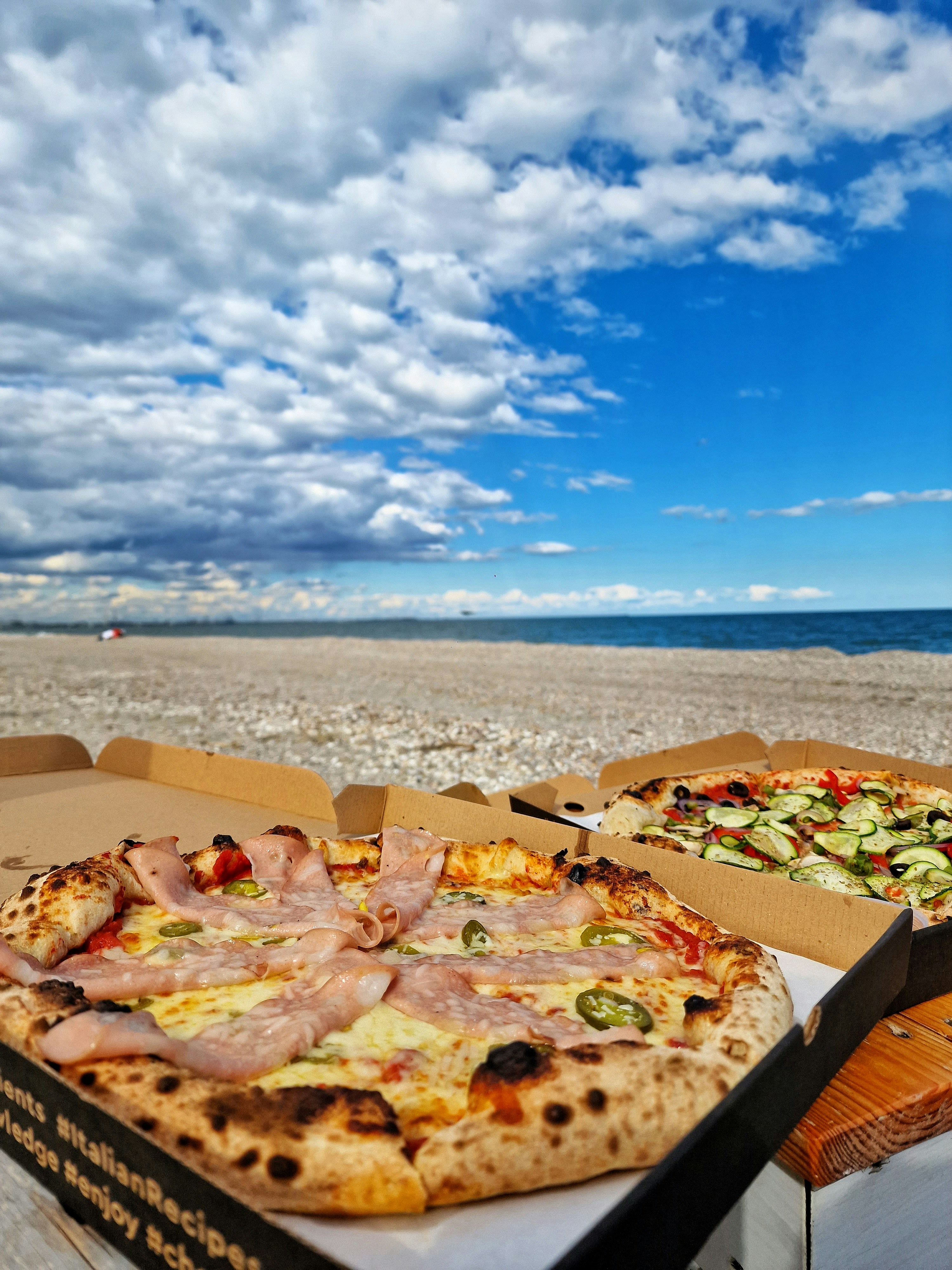 A scenic beach setting with two pizzas in open boxes on a wooden surface. The foreground pizza has slices of ham and green peppers on a crispy crust. The background shows another pizza with a variety of vegetables, including cucumbers and olives. The sky is partly cloudy with patches of bright blue, and the ocean is calm beyond the sandy shore.
