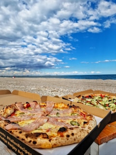 A scenic beach setting with two pizzas in open boxes on a wooden surface. The foreground pizza has slices of ham and green peppers on a crispy crust. The background shows another pizza with a variety of vegetables, including cucumbers and olives. The sky is partly cloudy with patches of bright blue, and the ocean is calm beyond the sandy shore.