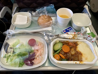 An airline meal tray featuring a main dish with beef stew, mashed potatoes, and vegetables such as carrots and cucumber. A side salad with lettuce, shrimp, and tomato is present alongside packaged butter. A roll wrapped in plastic and a cup of orange juice are included, with utensils and a napkin in a sealed wrapper.