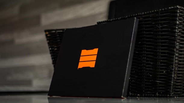 A black box featuring an orange emblem with three horizontal bars is positioned in front of a dark wicker basket. The background consists of a gray, textured wall.