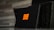 A black box featuring an orange emblem with three horizontal bars is positioned in front of a dark wicker basket. The background consists of a gray, textured wall.