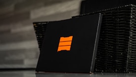 A black box featuring an orange emblem with three horizontal bars is positioned in front of a dark wicker basket. The background consists of a gray, textured wall.