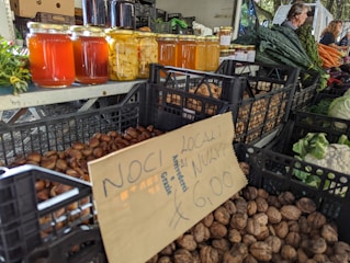 A market stall displays various jars of honey and preserved goods, alongside baskets filled with nuts and vegetables such as kale, carrots, and cauliflower. The setting suggests a busy farmers' market with people in the background organizing produce.