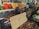 A market stall displays various jars of honey and preserved goods, alongside baskets filled with nuts and vegetables such as kale, carrots, and cauliflower. The setting suggests a busy farmers' market with people in the background organizing produce.