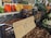 A market stall displays various jars of honey and preserved goods, alongside baskets filled with nuts and vegetables such as kale, carrots, and cauliflower. The setting suggests a busy farmers' market with people in the background organizing produce.