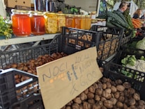 A market stall displays various jars of honey and preserved goods, alongside baskets filled with nuts and vegetables such as kale, carrots, and cauliflower. The setting suggests a busy farmers' market with people in the background organizing produce.