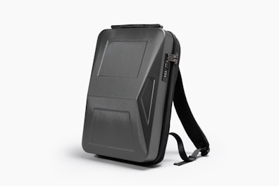 A sleek, black, hard-shell backpack with an ergonomic design. The front panel features distinct geometric patterns and a combination lock. The backpack has adjustable shoulder straps and a streamlined appearance, suitable for modern, urban settings.