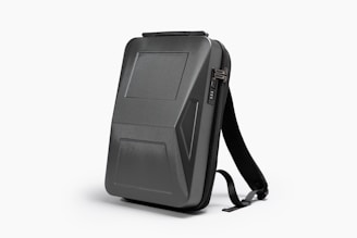 A sleek, black, hard-shell backpack with an ergonomic design. The front panel features distinct geometric patterns and a combination lock. The backpack has adjustable shoulder straps and a streamlined appearance, suitable for modern, urban settings.
