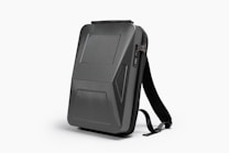 A sleek, black, hard-shell backpack with an ergonomic design. The front panel features distinct geometric patterns and a combination lock. The backpack has adjustable shoulder straps and a streamlined appearance, suitable for modern, urban settings.