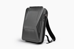 A sleek, black, hard-shell backpack with an ergonomic design. The front panel features distinct geometric patterns and a combination lock. The backpack has adjustable shoulder straps and a streamlined appearance, suitable for modern, urban settings.