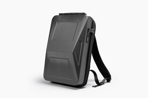 A sleek, black, hard-shell backpack with an ergonomic design. The front panel features distinct geometric patterns and a combination lock. The backpack has adjustable shoulder straps and a streamlined appearance, suitable for modern, urban settings.