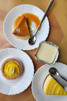 A collection of desserts is arranged on plates. There is a caramel flan on one plate with a spoon beside it, a small yellow frosted cupcake with a decorative lemon slice, a small tin container with a creamy dessert, and a slice of cheesecake with a yellow topping. Each dessert is presented on a separate plate with a silver spoon.