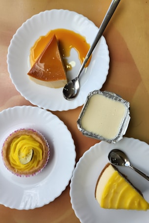 A collection of desserts is arranged on plates. There is a caramel flan on one plate with a spoon beside it, a small yellow frosted cupcake with a decorative lemon slice, a small tin container with a creamy dessert, and a slice of cheesecake with a yellow topping. Each dessert is presented on a separate plate with a silver spoon.