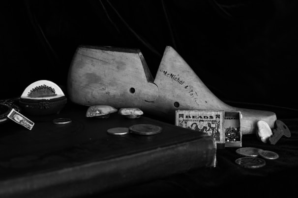 A black and white photograph featuring various antique and vintage objects arranged artfully. Prominent items include a wooden shoe form marked with 'McNichol & Taylor,' small boxes, some containing beads, coins scattered on a dark surface, and a decorative item that resembles a half kiwi. The setup conveys a sense of nostalgia and history.