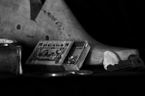 The image features a vintage setting with a wooden box labeled 'Beads' partially open, revealing colorful beads inside. The background includes a wooden object with text inscribed on it, possibly a shoe mold. In front of the box is a smooth stone and a large key. The overall composition is in black and white, lending a classic and nostalgic feel.