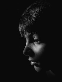 A black and white portrait featuring a person's face in profile with strong light emphasizing the textures and contours. The background is dark, highlighting the illuminated side of the face, especially the eye and lips.