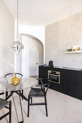 A modern kitchen interior features a minimalist design with light wooden flooring and a sleek black cabinet wall. The kitchen includes built-in appliances such as an oven and a counter with a coffee maker. A round glass dining table with black chairs is positioned nearby, and a metallic pendant light fixture hangs above. Decor elements include a yellow vase with ornamental grasses on the table and a wall shelf with fruit and a small plant.