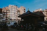 A traditional building with a dark tiled roof stands in front of a backdrop of residential apartment blocks. Numerous motorcycles are parked in the foreground, while a few cars are visible. The sky is clear, adding a sense of openness to the urban scene.