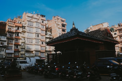 A traditional building with a dark tiled roof stands in front of a backdrop of residential apartment blocks. Numerous motorcycles are parked in the foreground, while a few cars are visible. The sky is clear, adding a sense of openness to the urban scene.