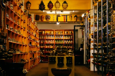 A shop interior filled with various cultural and decorative items, including masks, statues, and bowls displayed on shelves and tables. The shelves are organized with items displayed in an orderly fashion, and there is dim lighting creating a cozy atmosphere.