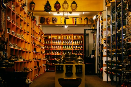 A shop interior filled with various cultural and decorative items, including masks, statues, and bowls displayed on shelves and tables. The shelves are organized with items displayed in an orderly fashion, and there is dim lighting creating a cozy atmosphere.