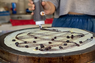 A person applies chocolate sauce in a zigzag pattern on a large, thin crepe while it's on a cooking surface. The crepe is topped with small dried fruits, likely raisins. The setting appears to be a kitchen or food stall with various utensils and a red cloth in the background.
