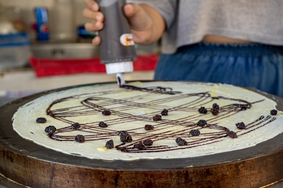 A person applies chocolate sauce in a zigzag pattern on a large, thin crepe while it's on a cooking surface. The crepe is topped with small dried fruits, likely raisins. The setting appears to be a kitchen or food stall with various utensils and a red cloth in the background.