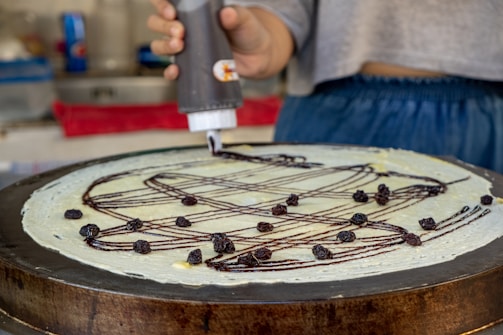 A person applies chocolate sauce in a zigzag pattern on a large, thin crepe while it's on a cooking surface. The crepe is topped with small dried fruits, likely raisins. The setting appears to be a kitchen or food stall with various utensils and a red cloth in the background.