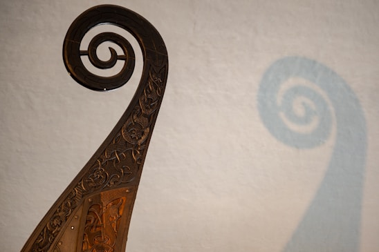 An intricately carved wooden spiral feature, reminiscent of a ship’s prow or ornamental decoration, casts a shadow on a plain backdrop. The carvings display detailed and flowing Celtic knot patterns.