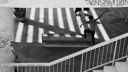 A black and white photograph of a person skateboarding in an urban environment with graffiti on the walls. The scene is lit by striped shadow patterns likely cast by overhead structures, creating an artistic effect on the concrete floor. A rail is visible in the foreground, and there are chairs in the background.