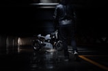 A person dressed in a black leather jacket and dark pants walks through a dimly lit urban environment, holding a motorcycle helmet. In the background, a sleek, modern motorcycle is parked, its metallic surface reflecting the limited light.