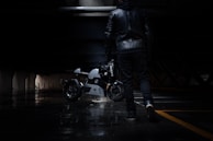 A person dressed in a black leather jacket and dark pants walks through a dimly lit urban environment, holding a motorcycle helmet. In the background, a sleek, modern motorcycle is parked, its metallic surface reflecting the limited light.