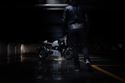A person dressed in a black leather jacket and dark pants walks through a dimly lit urban environment, holding a motorcycle helmet. In the background, a sleek, modern motorcycle is parked, its metallic surface reflecting the limited light.