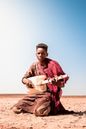 A person is seated on the ground in a desert-like environment, playing a traditional string instrument crafted from a plastic container. They are dressed in earthy-colored clothing with a red cloth draped over their shoulders. The background is an expansive clear blue sky.