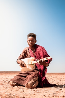 A person is seated on the ground in a desert-like environment, playing a traditional string instrument crafted from a plastic container. They are dressed in earthy-colored clothing with a red cloth draped over their shoulders. The background is an expansive clear blue sky.