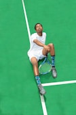 A person wearing a white sports outfit is sitting on a bright green tennis court. The individual has their eyes closed and appears relaxed. They are holding a tennis racket in one hand, and wearing gray sneakers and blue patterned socks.